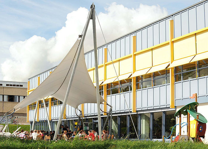 A large sunshade made of a metal frame and stable linen fabric stands next to a blue and yellow cafeteria building.